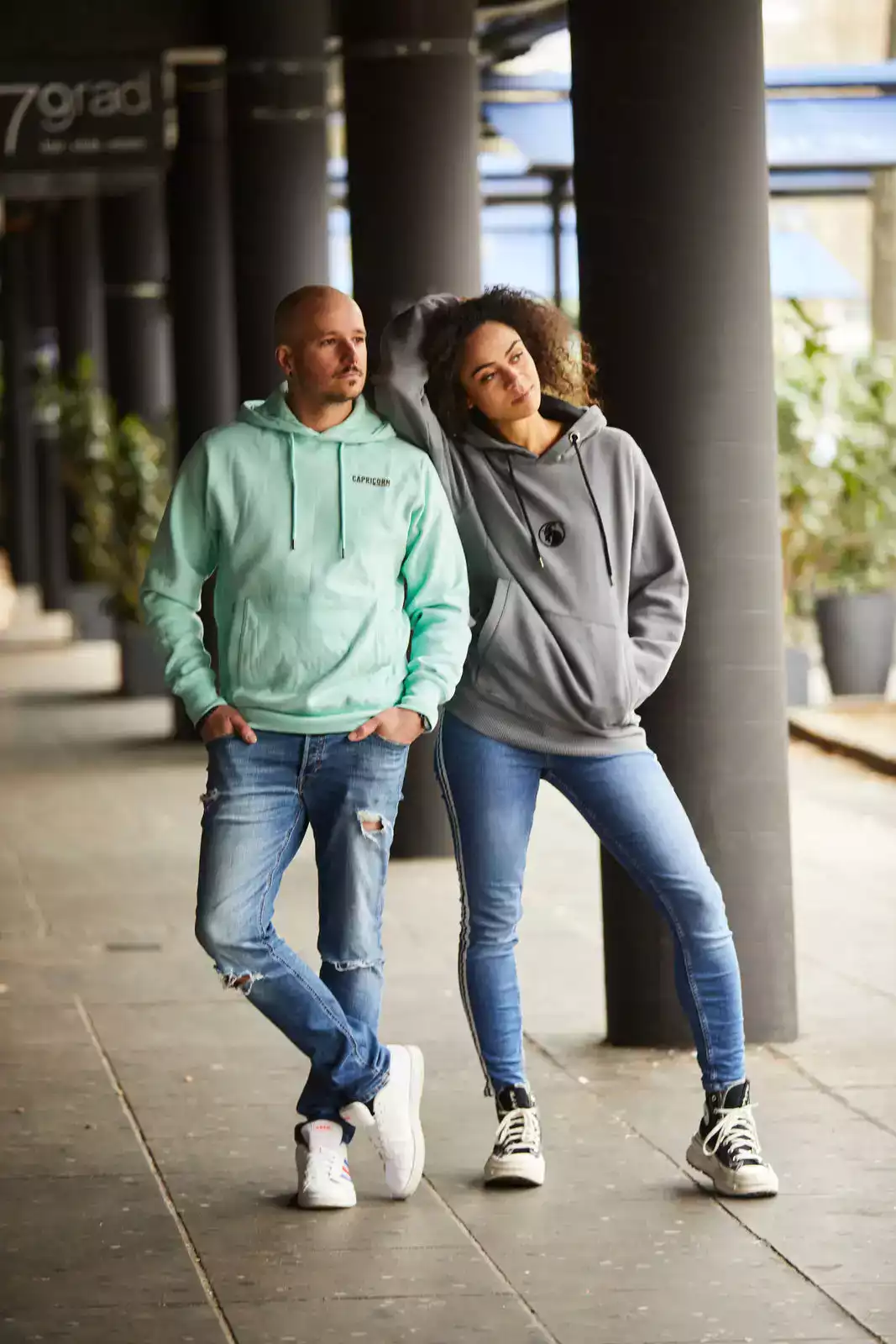 Männliches Model mit mintfarbenem Hoodie und weibliches Model mit grauem Hoodie stehend in Passage.