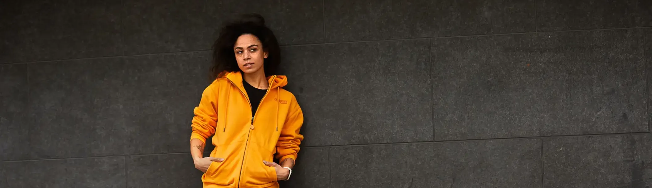 Weibliches Model mit orangefarbener Kapuzenjacke vor dunkler Wand