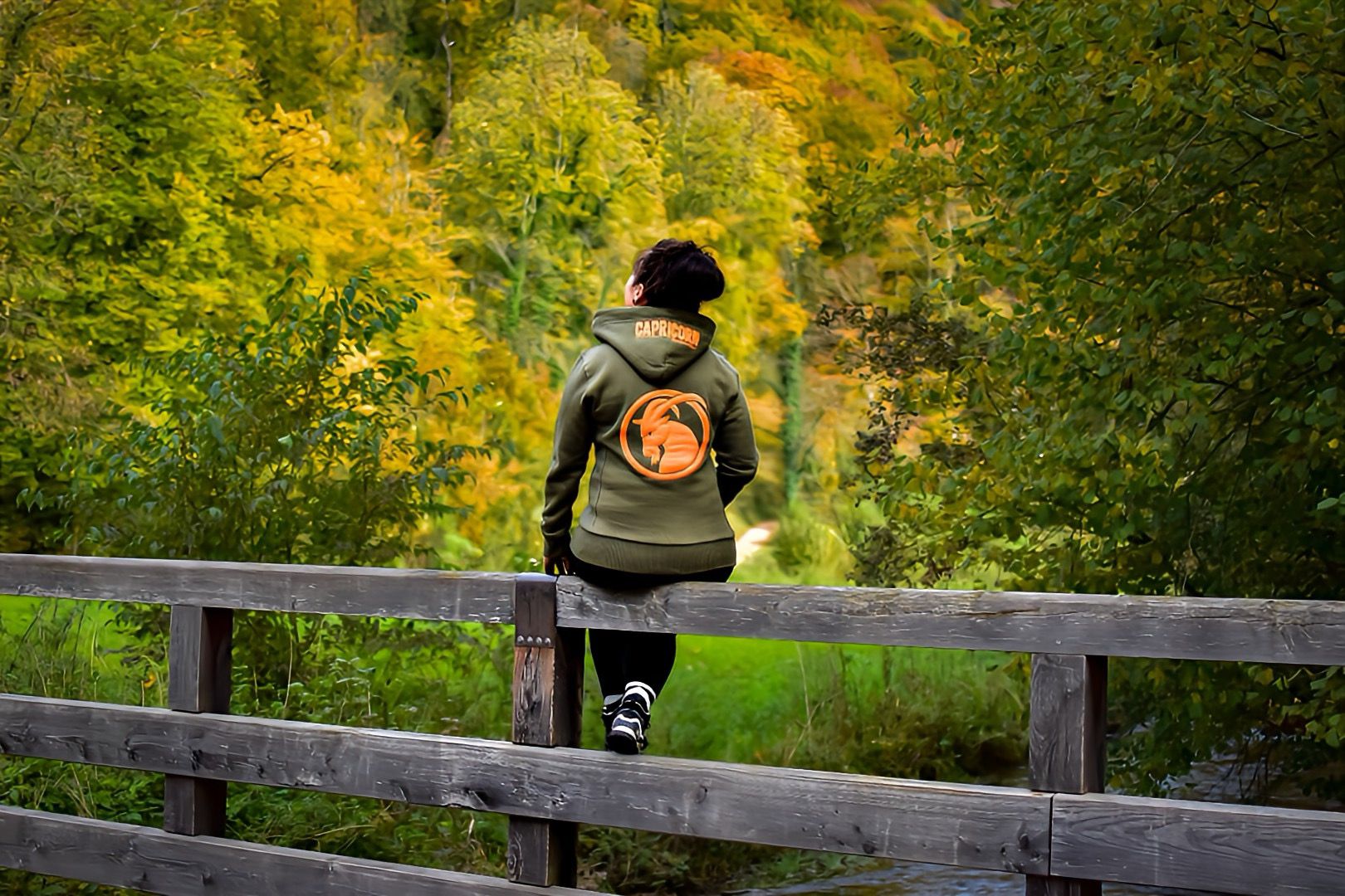Hoodie olivgrün mit orangefarbenem Steinbockkopf auf dem Rücken getragen von Frau auf Balken sitzend