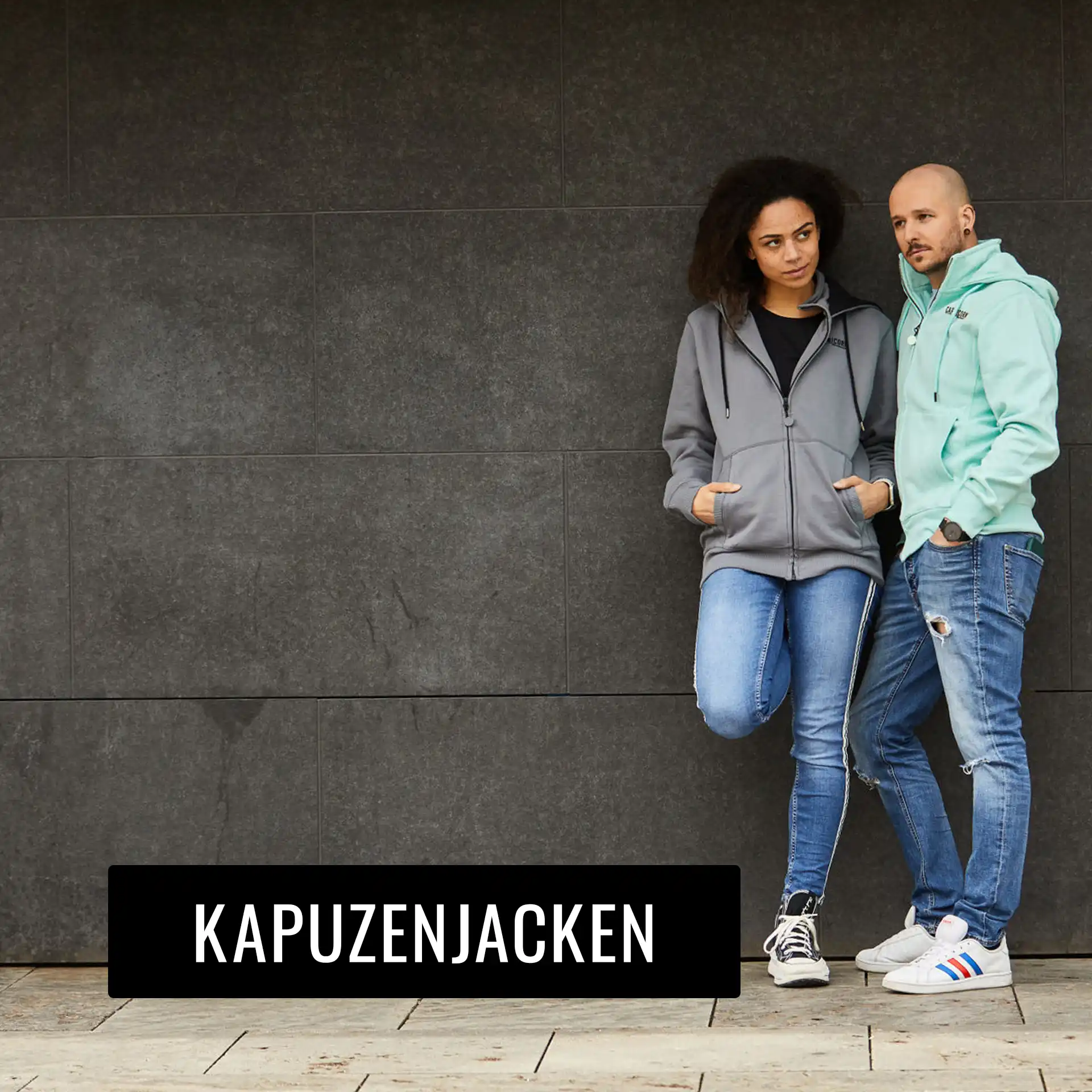 Frau mit grauer Kapuzenjacke und Mann mit mintfarbener kapuzenjacke vor dunkler Wand stehend.