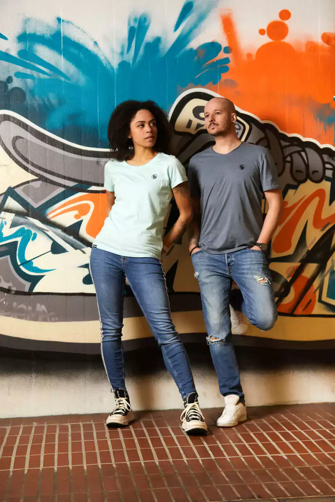 Frau mit mintfarbenem T-Shirt und Mann mit grauem T-Shirt vor Graffiti Wand stehend