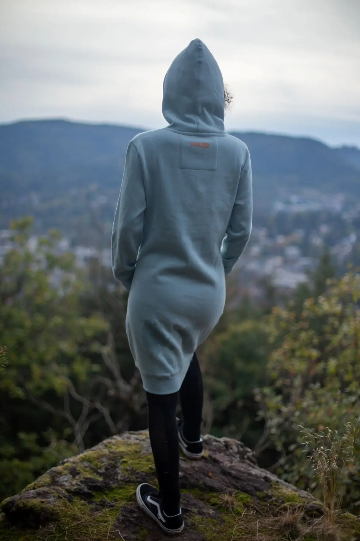 Model mit Sweatkleid hellblau auf Felsen stehen in die Ferne schauend. Rückansicht.