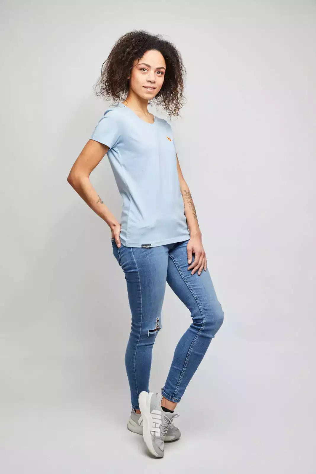 Frau trägt hellblaues T-Shirt mit orangefarbenem Stick vor weißer Wand