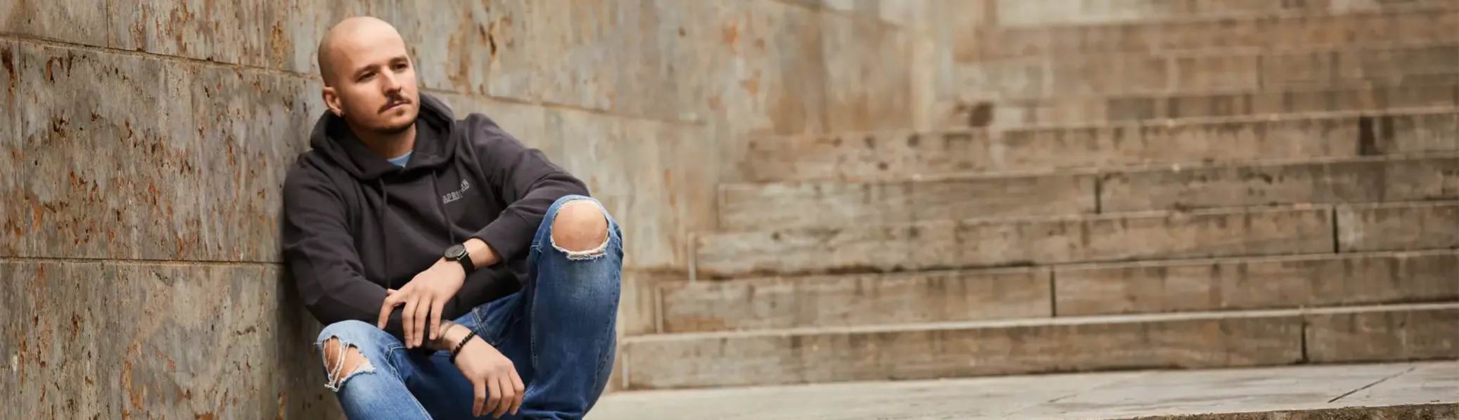 Banner Bild männliches Model sitzt auf Treppe und trägt dunkelgrauen Hoodie
