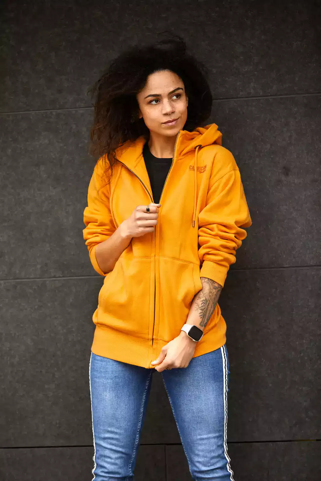 Weibliches Model trägt orangefarbene Kapuzenjacke vor dunkler Wand