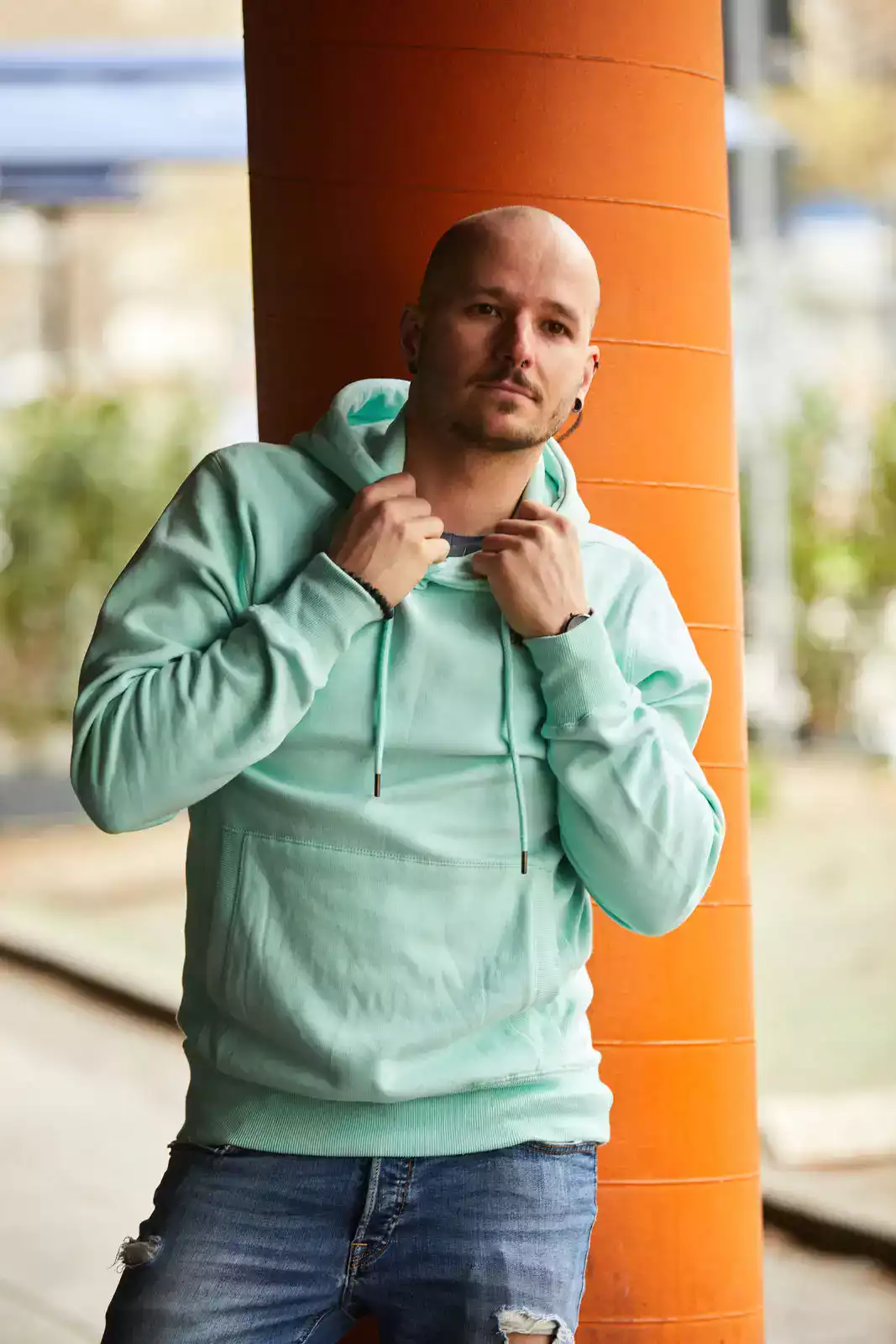 Männliches Model mit mintfarbenem Hoodie vor orangefarbener Säule stehend.